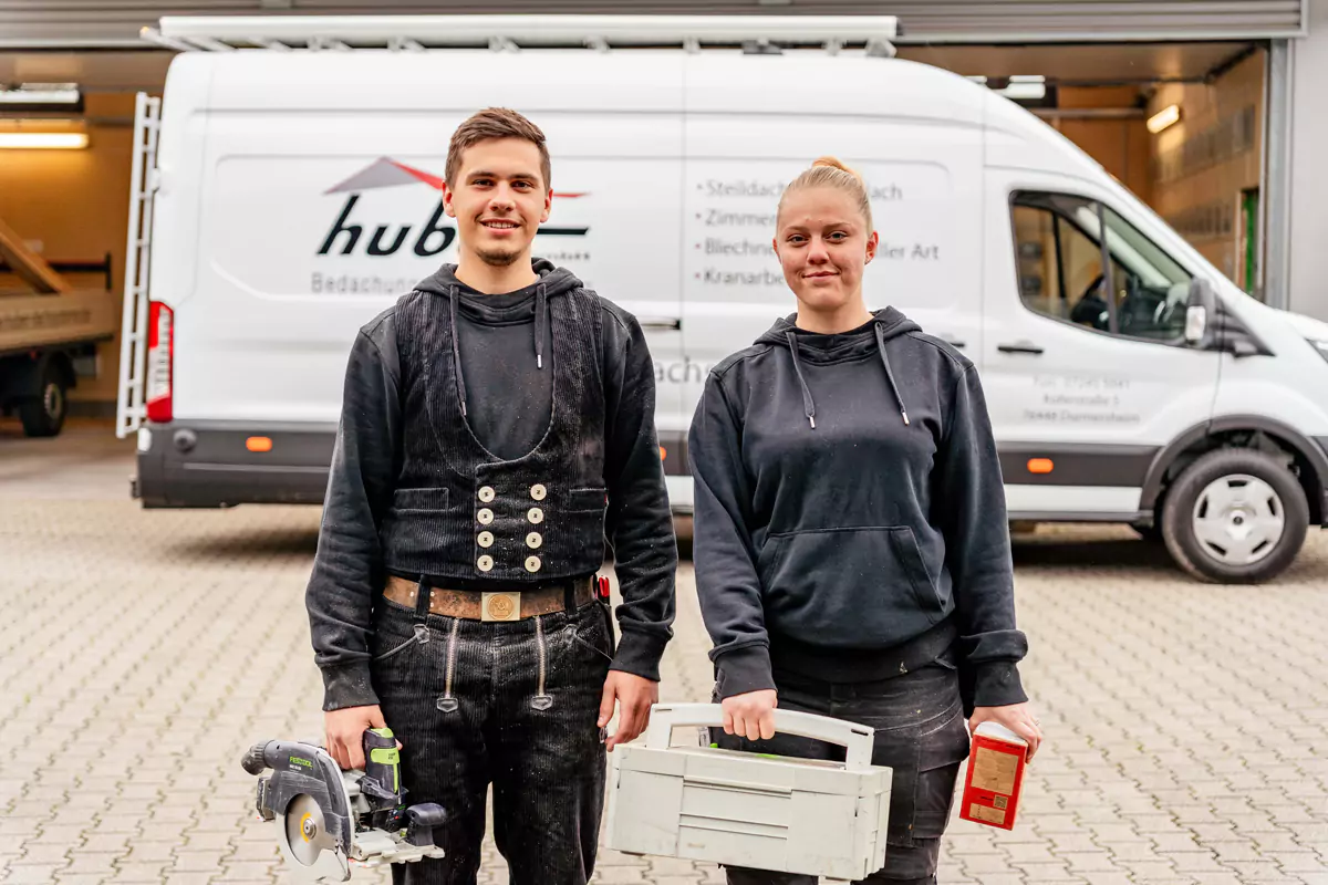 2 Dachdecker:innen mit Werkzeugkoffer und kleiner Kreissäge stehen vor einem Firmenauto