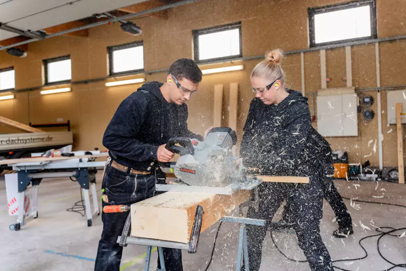 2 Dachdecker:innen sägen einen Holzbalken mit einer Kreissäge